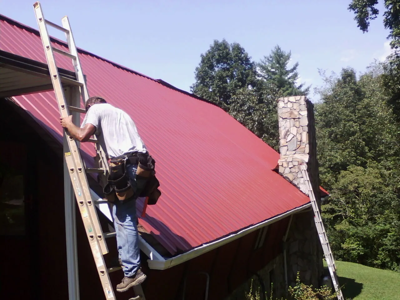 Roofer on a ladder beside a red metal roof and stone chimney — Soffit & Fascia Repair by Roofing Brothers in Industry